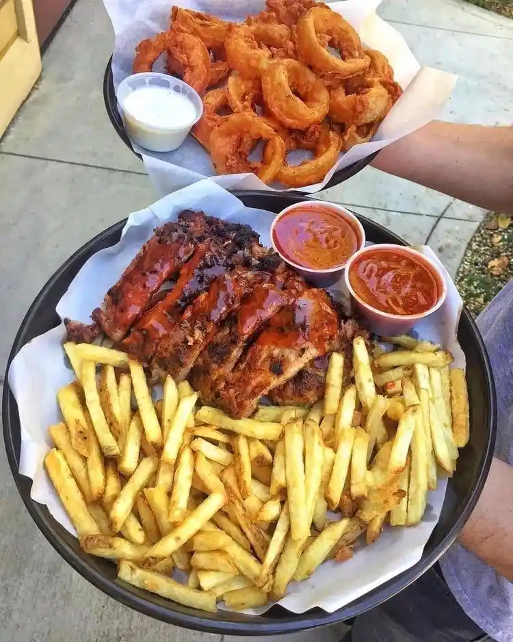 Two people hold up large plates of food outdoors. The foreground plate has barbecue ribs, french fries, and two small cups…