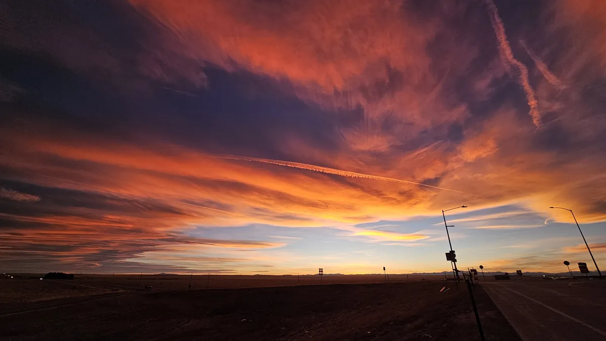 A dramatic, colorful sunset over a flat, open landscape with a road and streetlights visible in the foreground. The sky is…