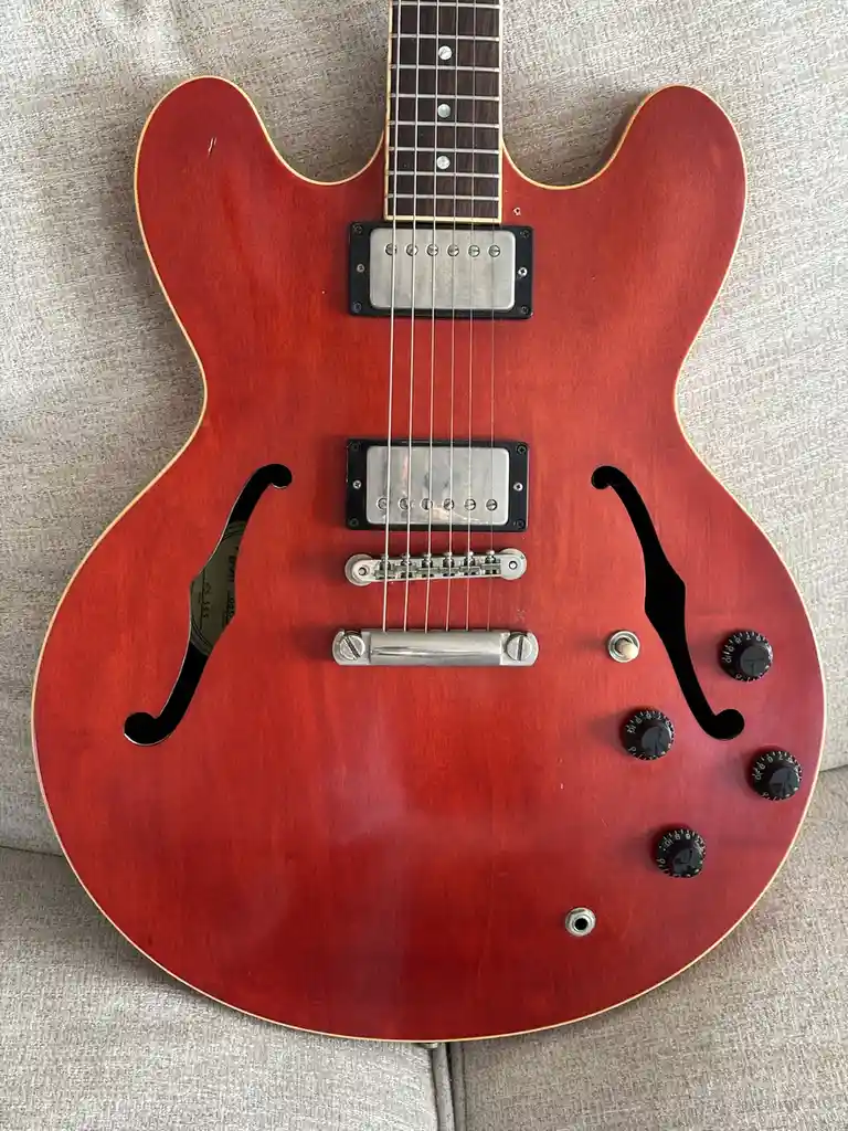 A red semi-hollow body electric guitar with f-holes, two humbucker pickups, and control knobs rests on a beige fabric…