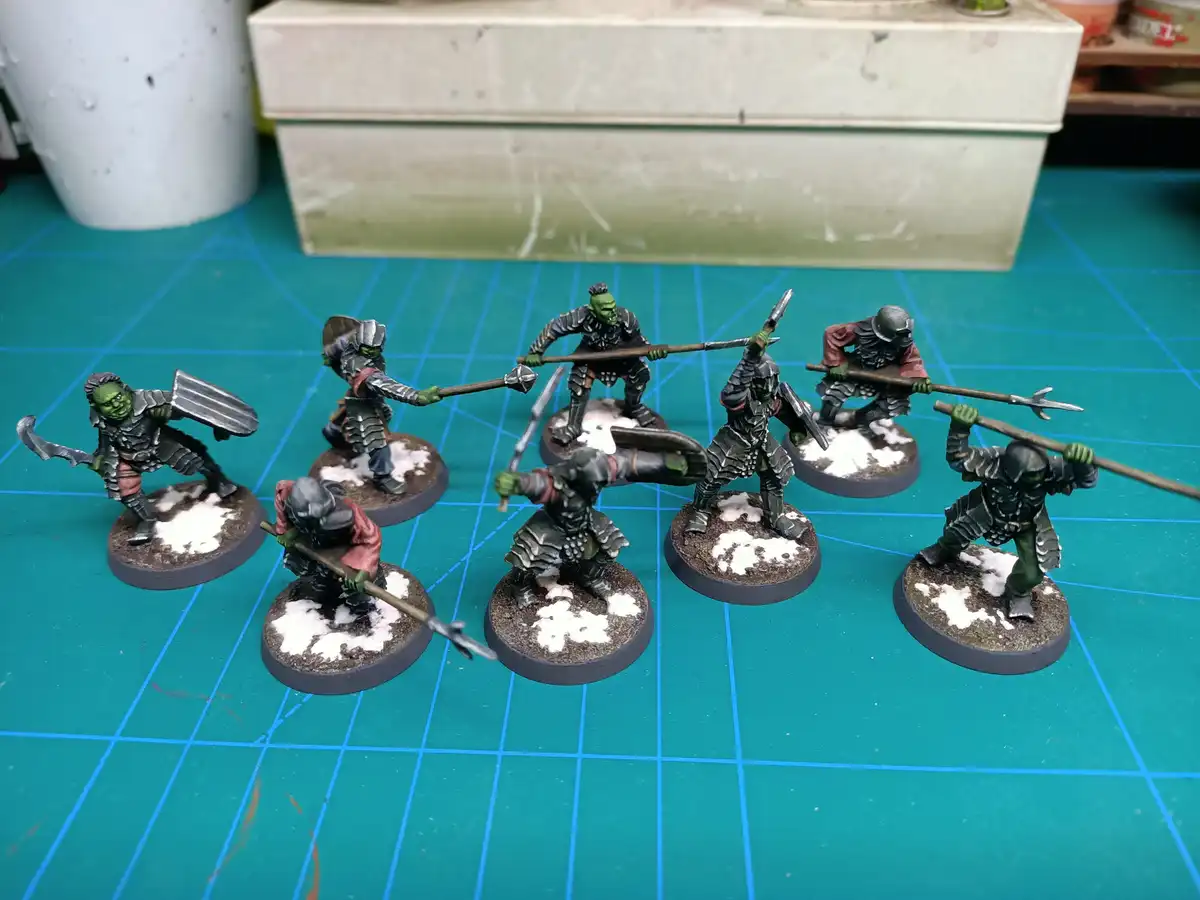 Seven painted miniature fantasy orcs in armor stand on circular bases with snow-like paint on them, arranged on a green grid…