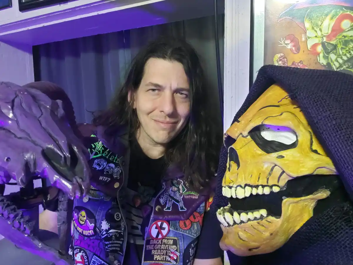 A man with long dark hair wearing a heavily patched denim jacket stands beside a large yellow skull mask with a purple hood…
