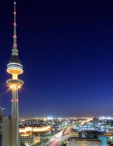 A nighttime photo of the illuminated Kuwait Tower against a dark blue sky, with city lights and traffic trails visible…
