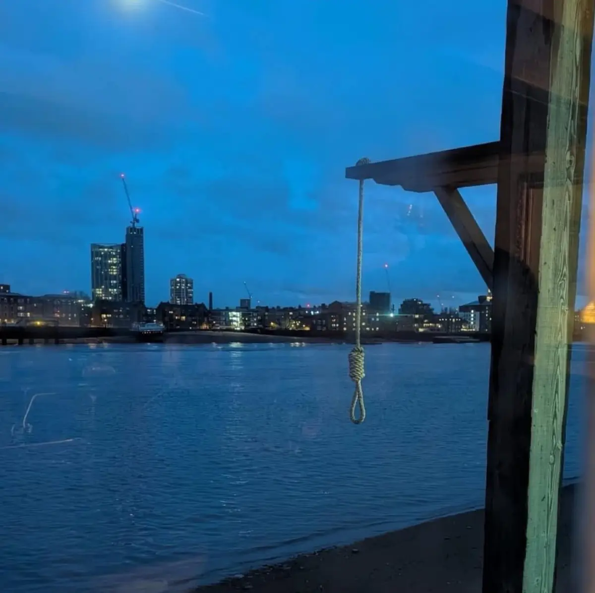 A photo taken from inside a building looking out at a city skyline at dusk, with a noose hanging from a wooden structure in…
