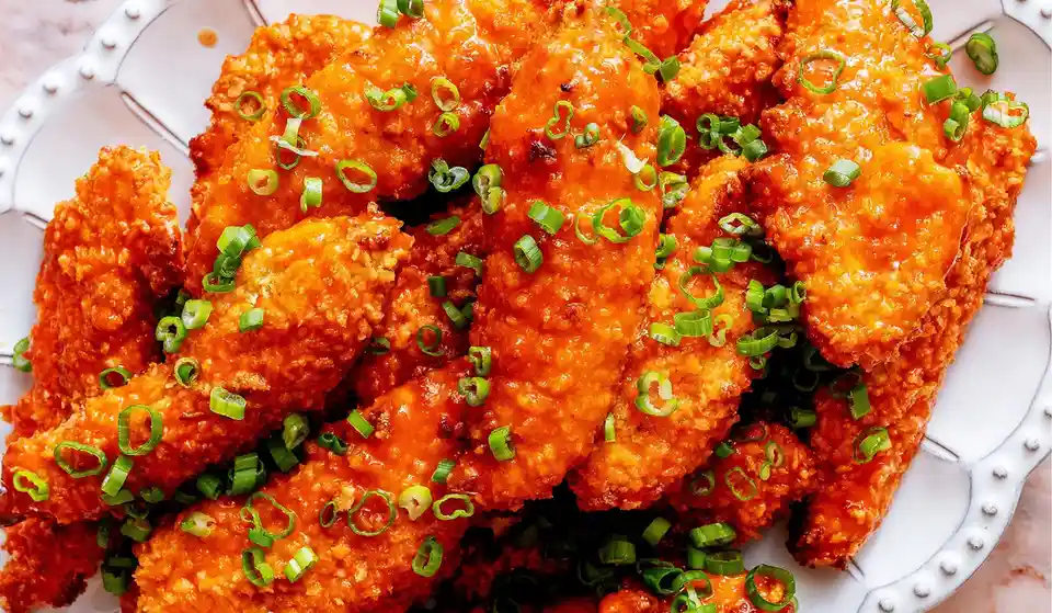 A close-up photo of crispy, orange buffalo sauce-covered chicken wings piled on a white decorative plate, garnished with…