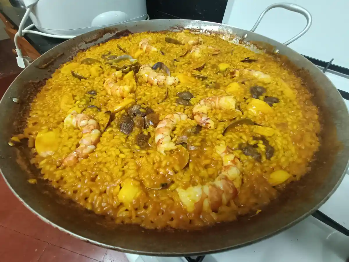 A large, shallow metal pan filled with a vibrant yellow rice dish, likely paella, topped with cooked shrimp, clams, and…