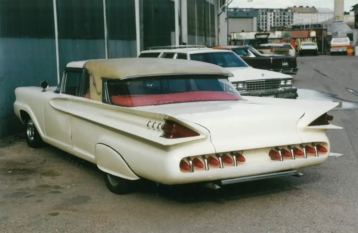 a white 1950s american car with a tan convertible top and red interior is parked in a lot, its rear features prominent…