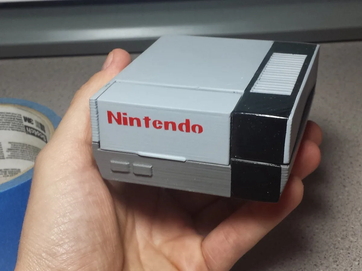 A hand holds a small, 3D-printed replica of a classic Nintendo Entertainment System console, featuring its iconic gray and…