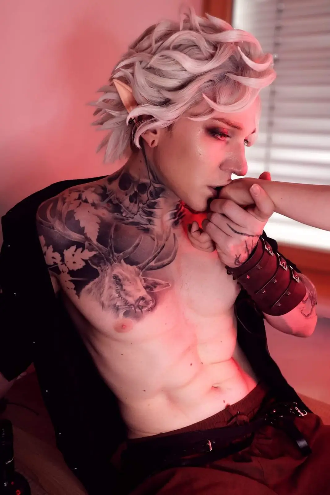 A person with pale blonde hair, pointed elf ears, and extensive tattoos including a large deer on their chest and skull…