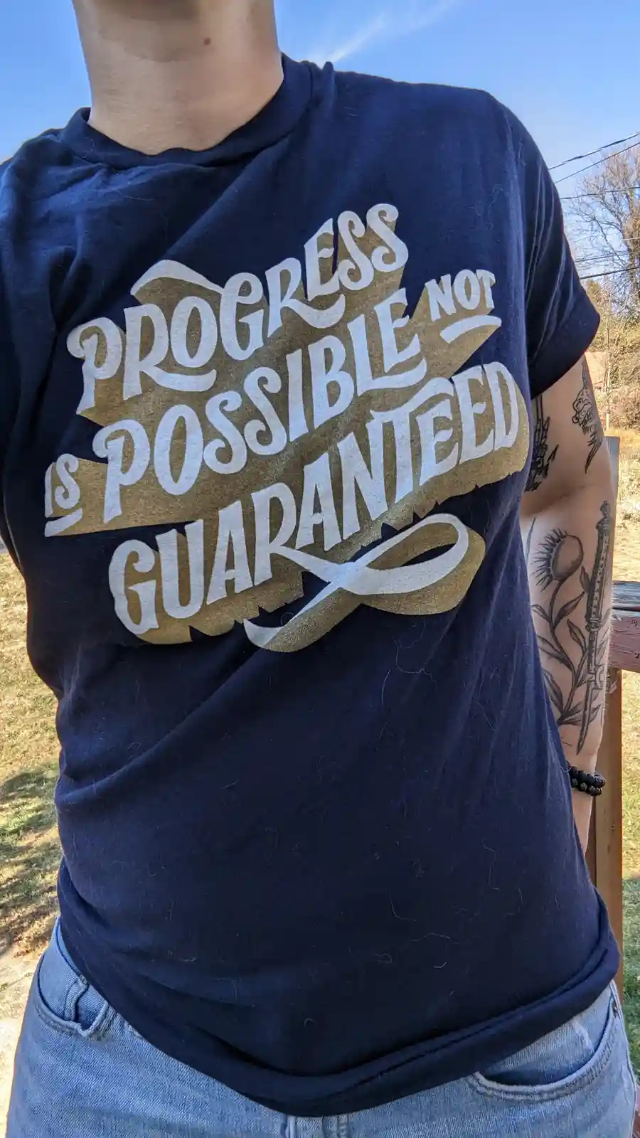 A person wearing a navy blue t-shirt with the phrase 'progress is possible not guaranteed' printed in white and gold…