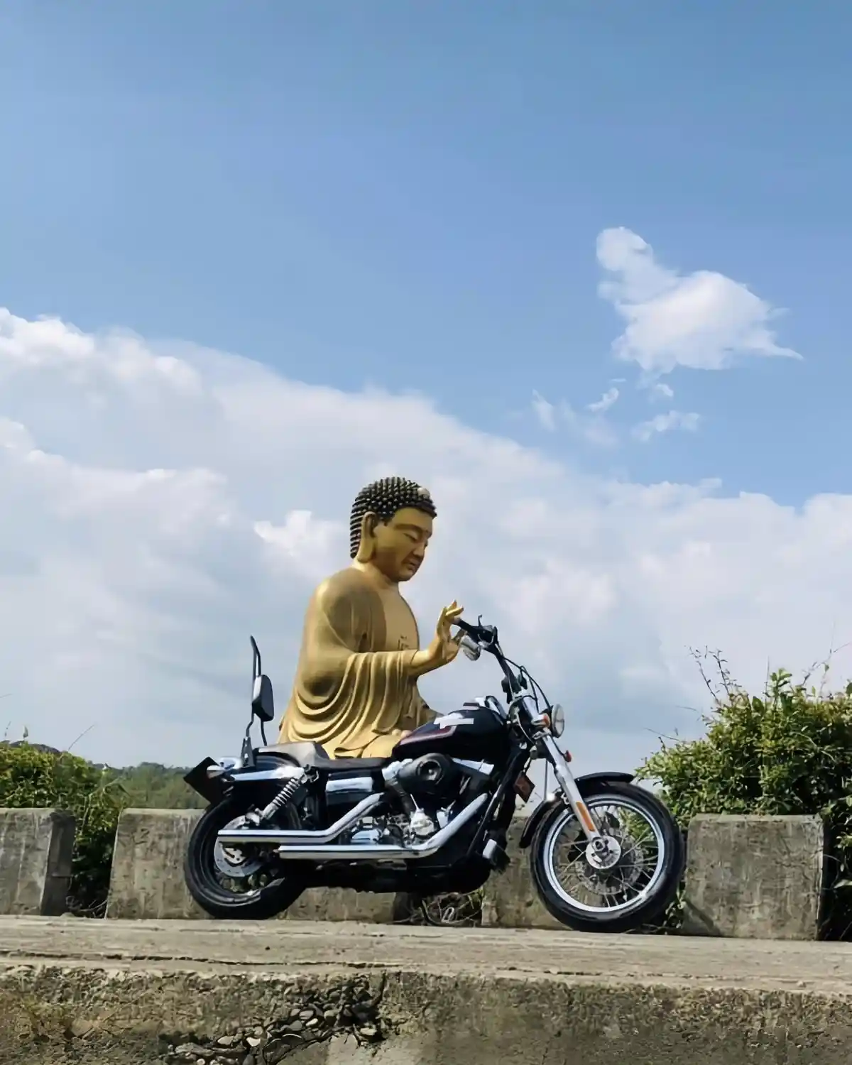 A large golden Buddha statue is seated on a black motorcycle, appearing to hold the handlebars, against a backdrop of blue…