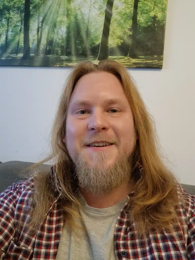 A man with long blonde hair and a beard, wearing a plaid shirt over a t-shirt, smiles at the camera while seated. Behind…