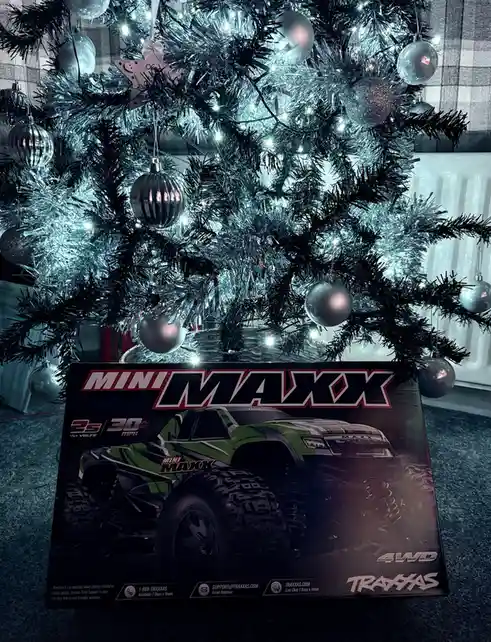 A box for a Traxxas Mini Maxx 4WD remote-controlled monster truck is placed on the floor in front of a decorated Christmas…