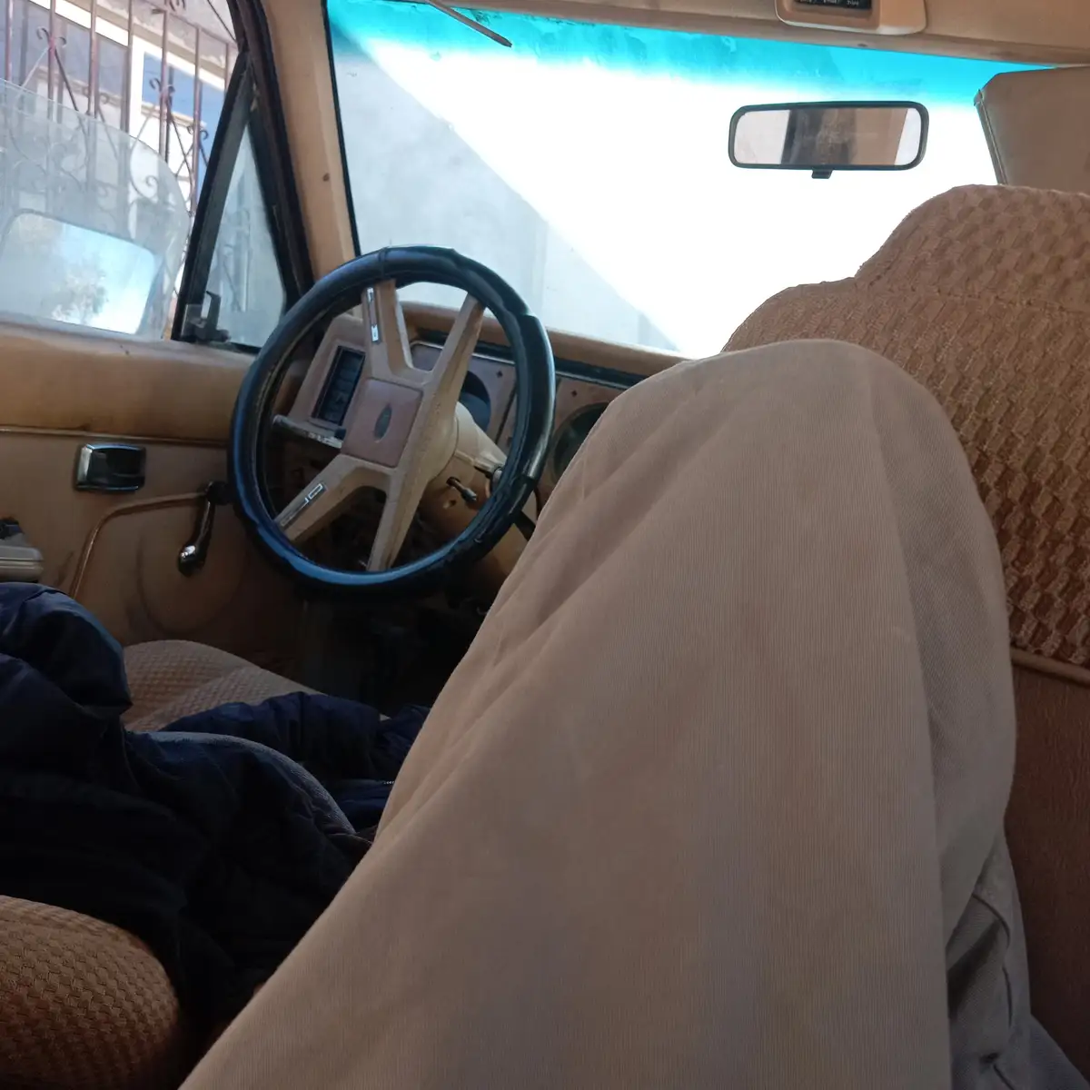 A first-person view from inside an old, beige car with a worn steering wheel and patterned seats, showing a person's leg in…