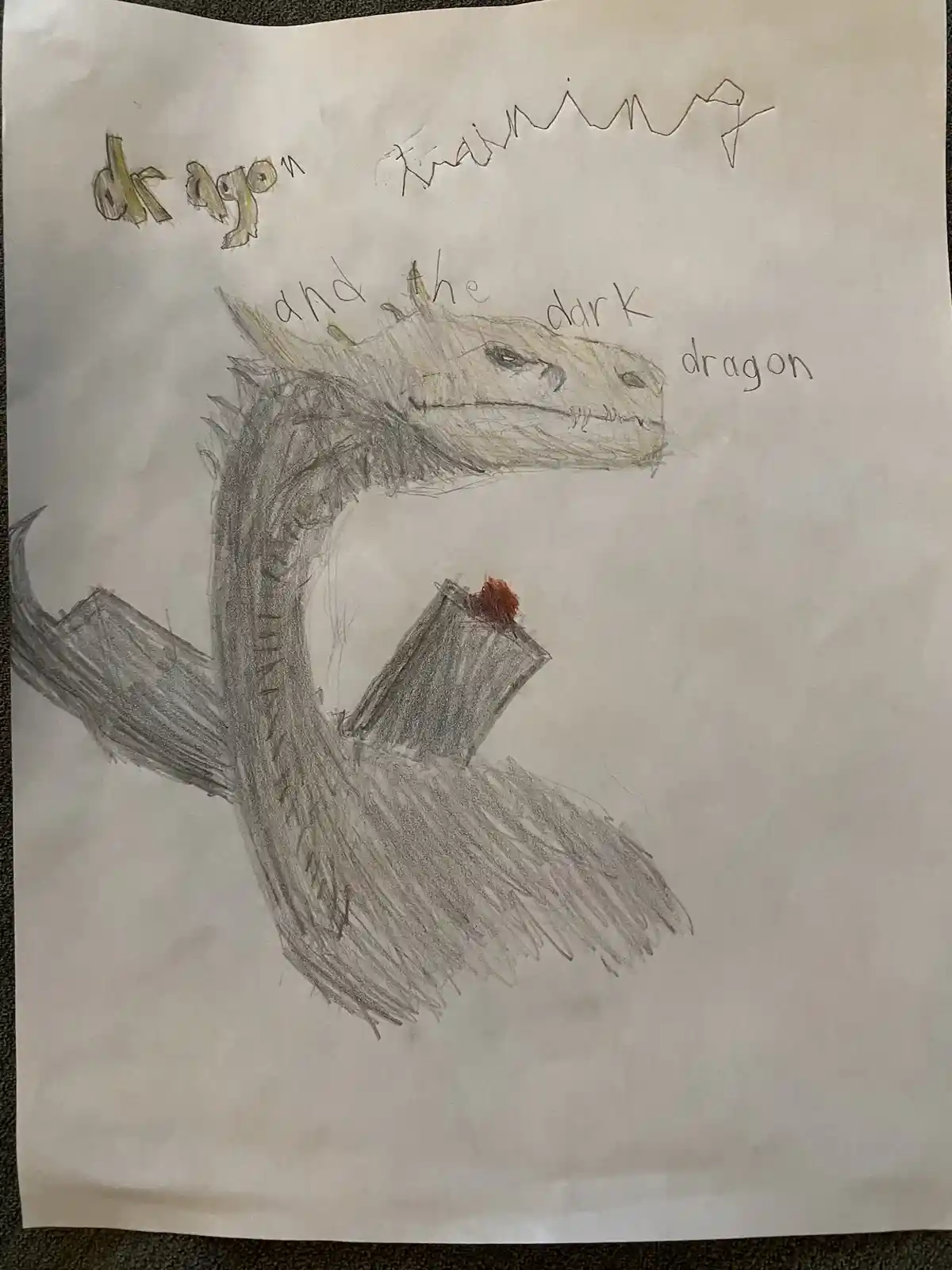 A child's crayon and pencil drawing shows a large, grey dragon with spiky scales, a long neck, and a small red flame on its…