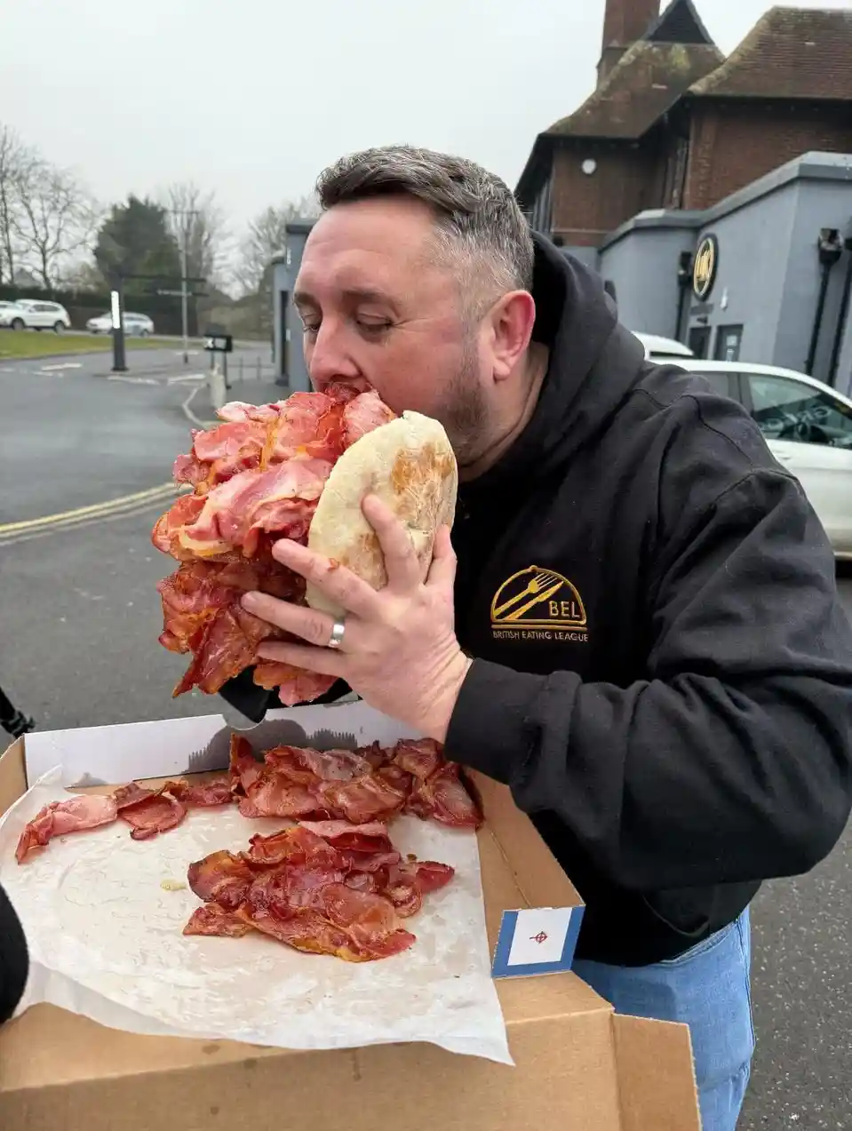 A man in a black hoodie with 'British Eating League' logo is eating a massive sandwich overflowing with bacon, held over a…