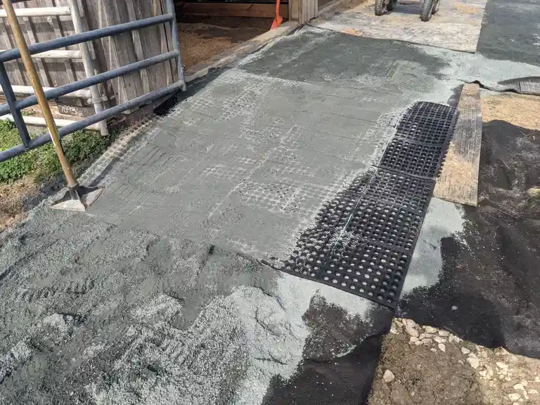 A freshly poured concrete surface at a construction site shows a distinct, uneven pattern where a black, perforated rubber…