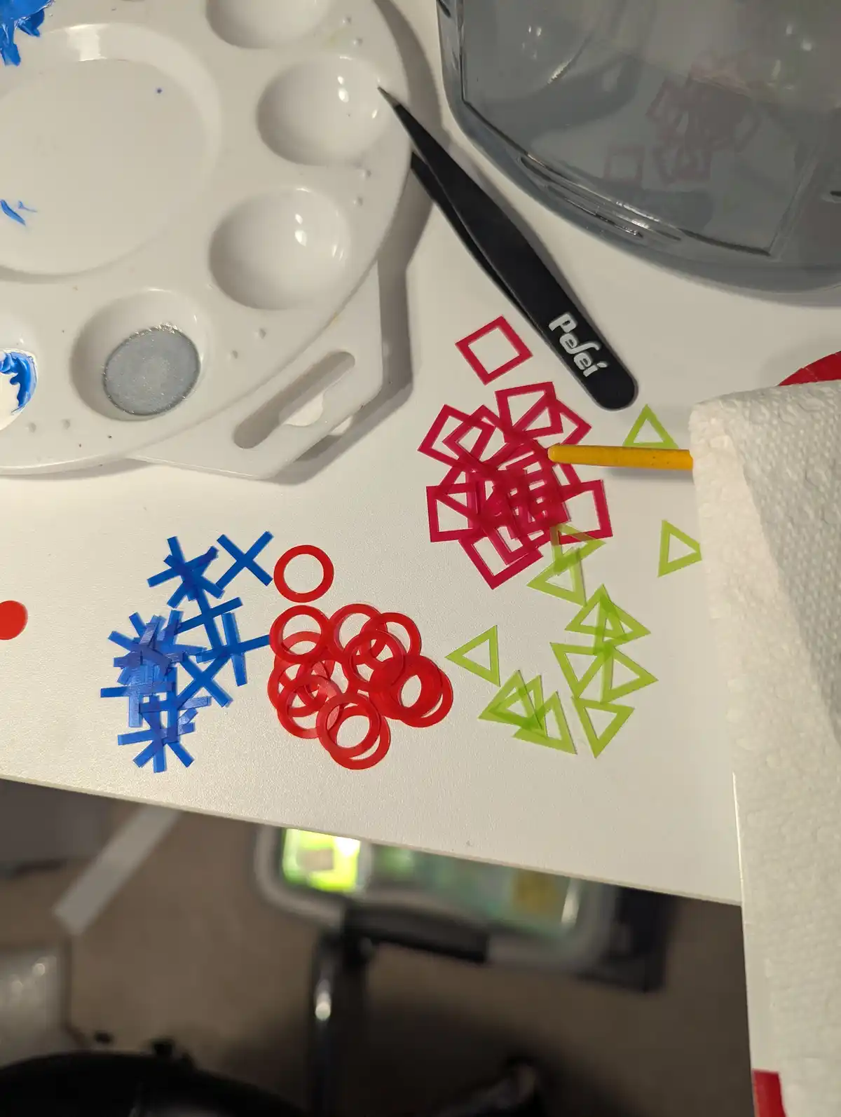 A close-up photo shows a white crafting surface with three groups of colorful rubber bands arranged in tic-tac-toe shapes…