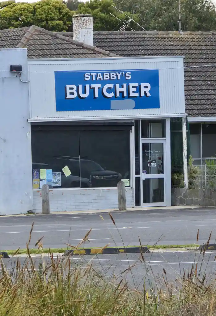 An exterior shot of a butcher shop named 'STABBY'S BUTCHER' with a blue sign. The shop's door has a 'Sorry CLOSED' sign on…