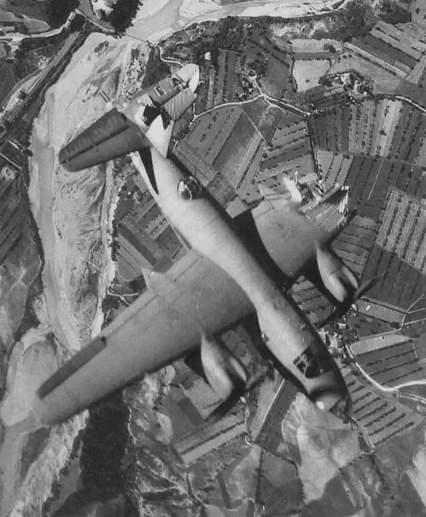 A black-and-white aerial photograph shows a military aircraft flying over patchwork farmland, viewed from directly above…
