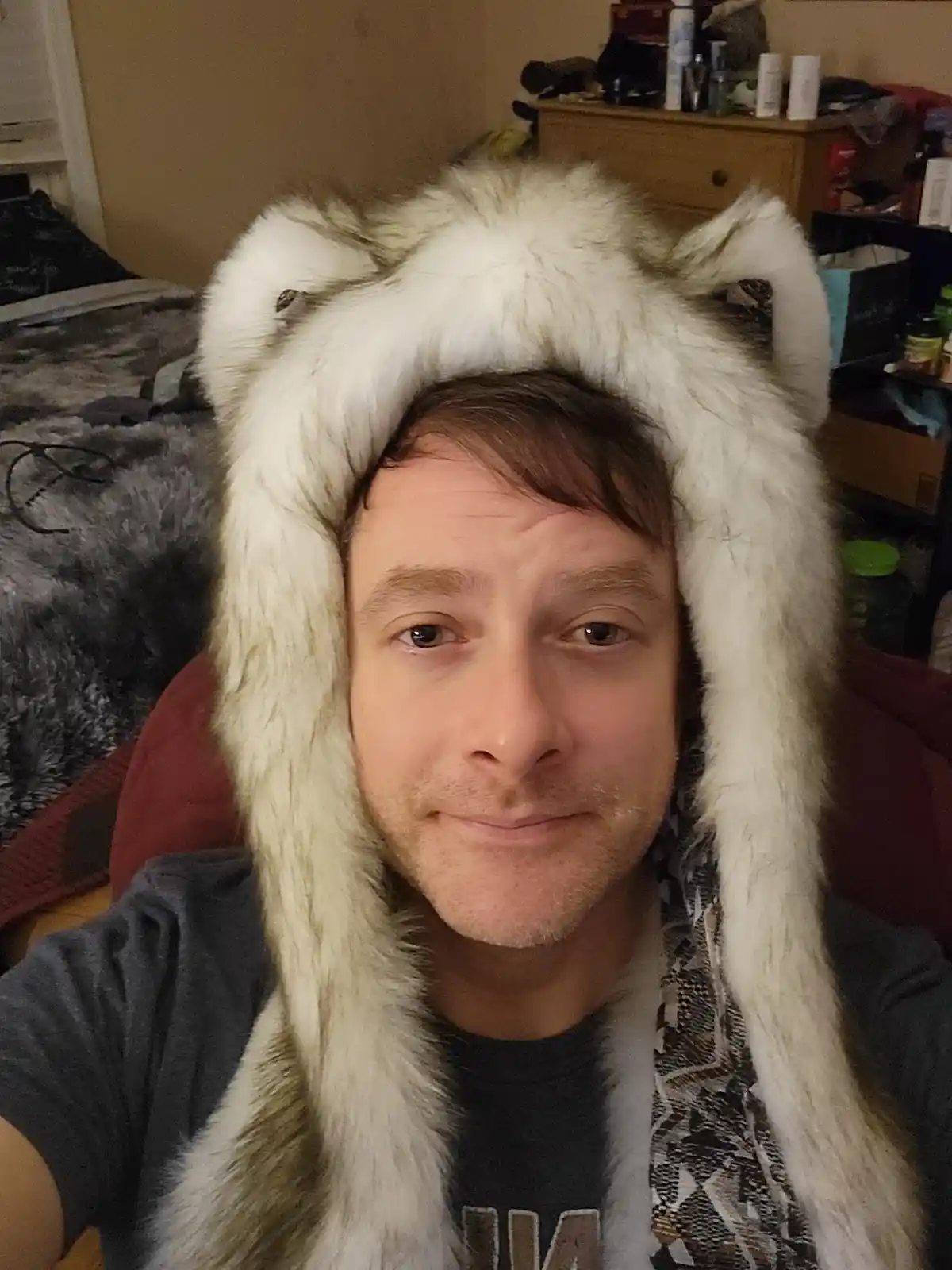 A man in a selfie wearing a large, fluffy white bear hat with ear flaps, looking directly at the camera with a subtle smirk…