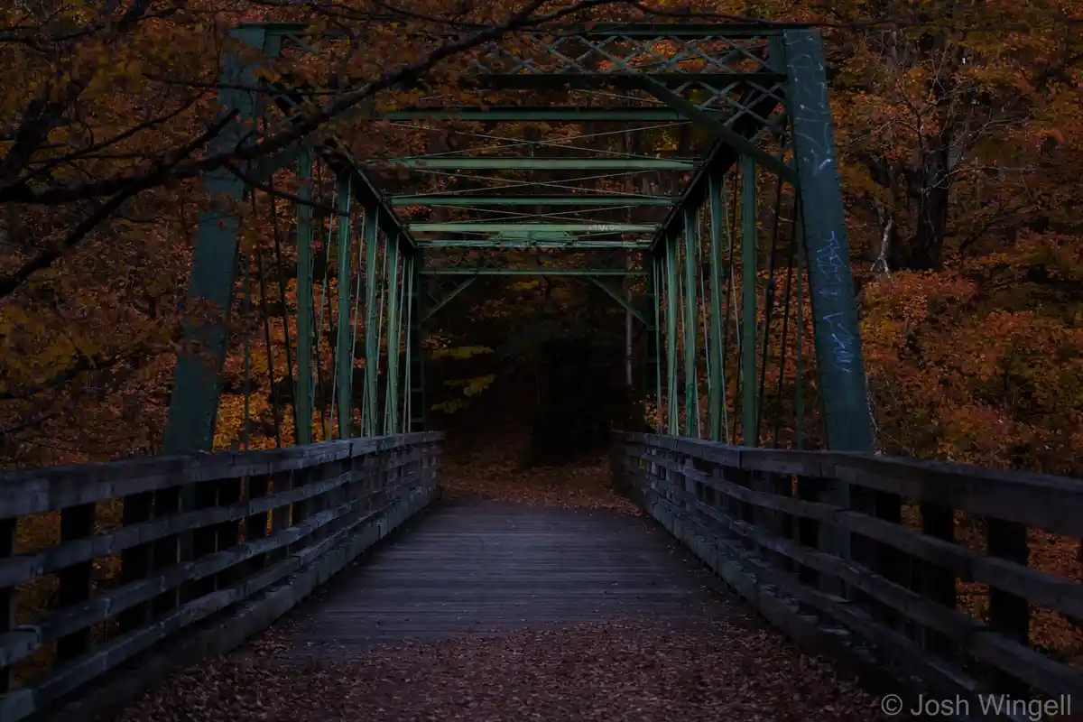 A dark, atmospheric photo of a green metal truss bridge with wooden planks and railings, surrounded by autumn trees with…