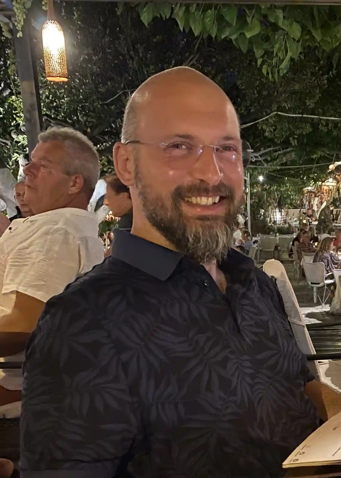 A bald man with glasses and a beard smiles while seated at an outdoor restaurant at night, wearing a dark patterned shirt…