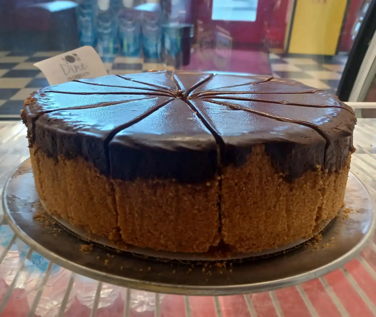 A round chocolate cake with a glossy chocolate glaze, pre-sliced into wedges, sits on a metal cake stand inside a glass…