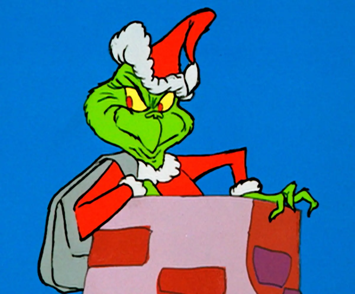 a green, grumpy cartoon character with yellow eyes and a red Santa hat with white trim peers over a red and pink brick wall…