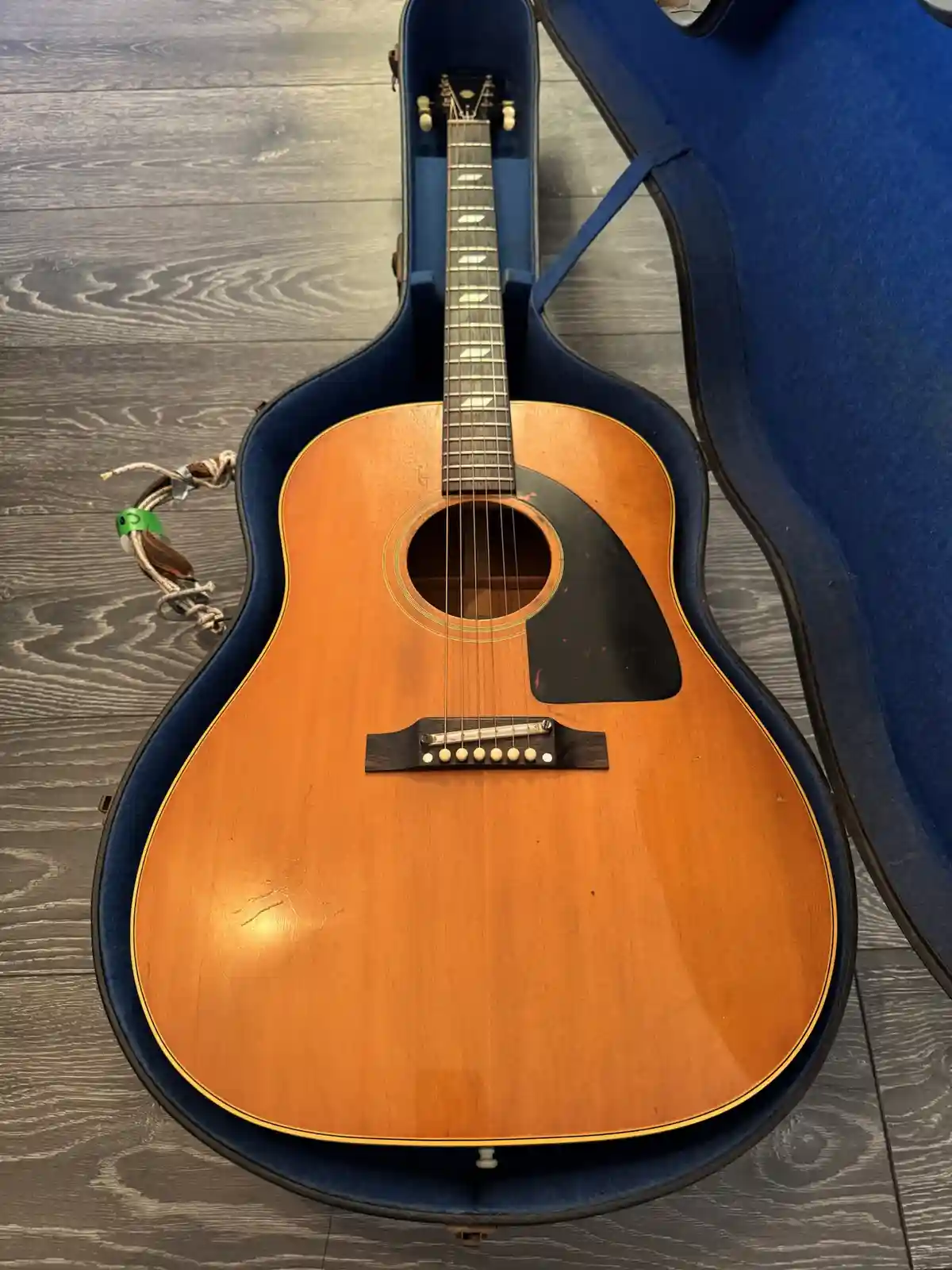 An acoustic guitar with a natural wood finish sits inside an open blue hardshell case on a gray wood floor. The guitar has a…