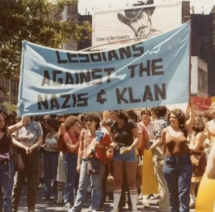 A vintage photograph shows a crowd of people, mostly women, marching in a street protest holding a large light blue banner…