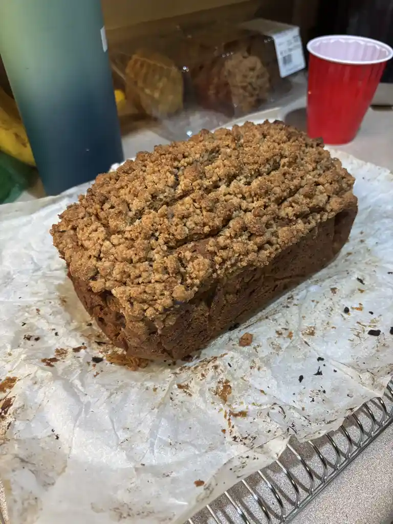 A freshly baked loaf of crumb-topped bread sits on parchment paper atop a cooling rack, with a red plastic cup and a blue…