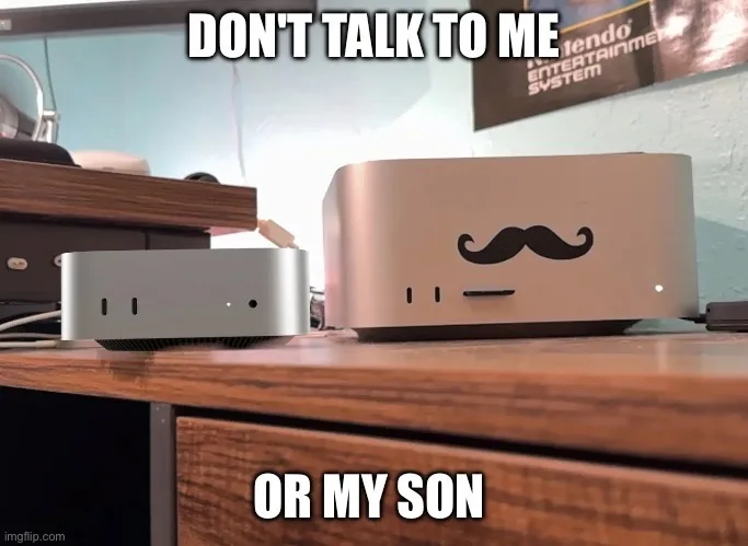 Two silver Apple Mac Mini computers sit side-by-side on a wooden desk. The larger one on the right has a black mustache…