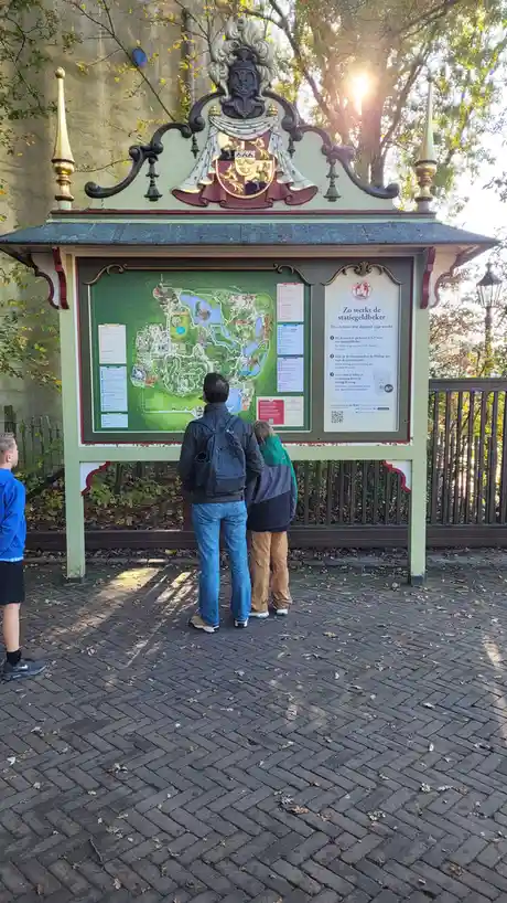 Three people, including a child, stand before a large ornate park map kiosk at Efteling, with the sun shining through trees…