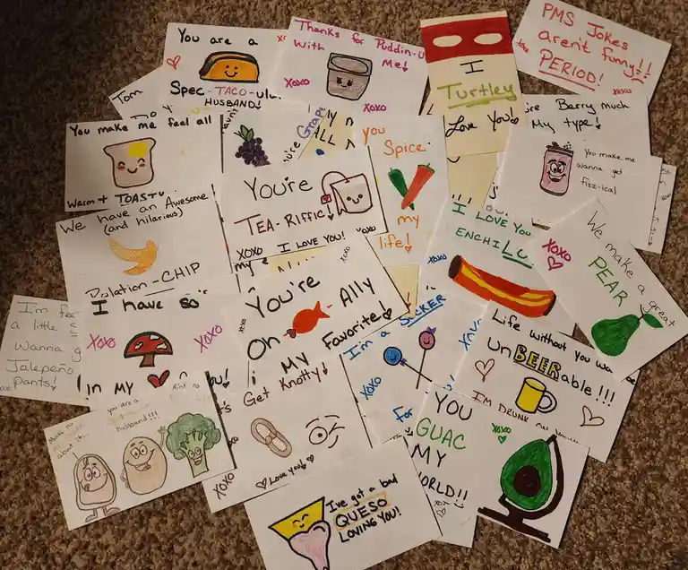 A pile of handmade, hand-drawn love notes on white paper scattered on a carpet, each featuring a cute cartoon food item and…