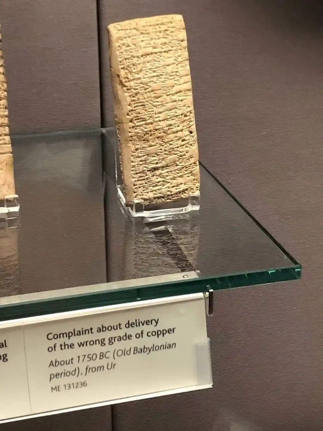 A museum display shows an ancient cuneiform tablet on a glass shelf, with a label describing it as a 'Complaint about…