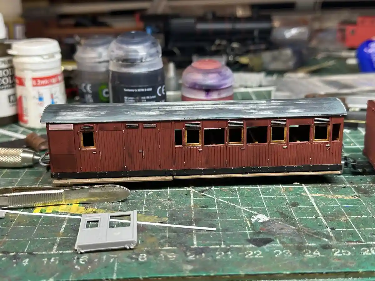 A close-up photo shows a detailed, reddish-brown model train passenger car with a weathered grey roof, sitting on a green…