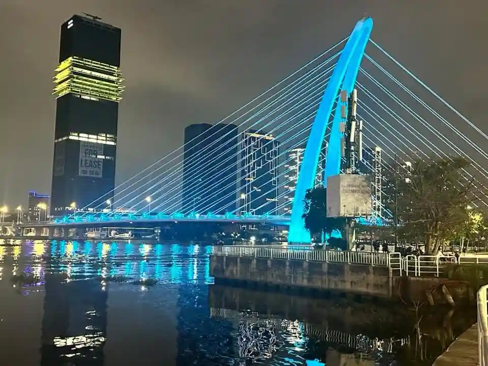 A nighttime cityscape featuring a brightly lit blue cable-stayed bridge over water, with modern skyscrapers in the…