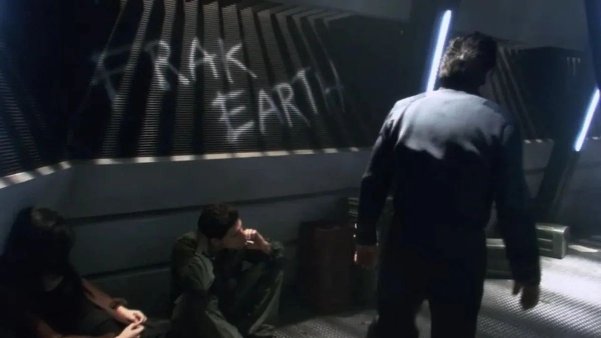 In a dimly lit sci-fi setting, two people sit dejectedly on the floor while a man walks away from them. Graffiti reading…