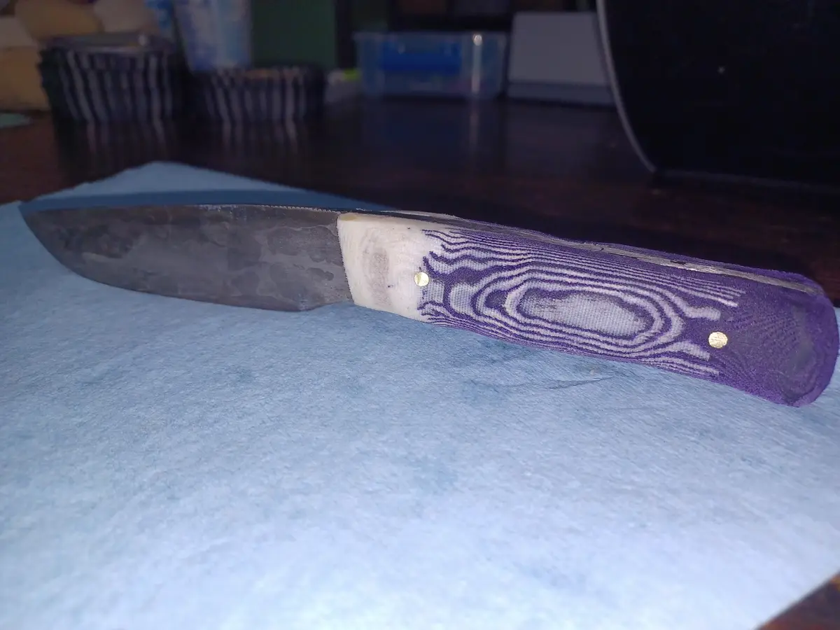 A close-up photo shows a handmade knife with a dark, textured blade and a purple handle featuring a wood-grain pattern…