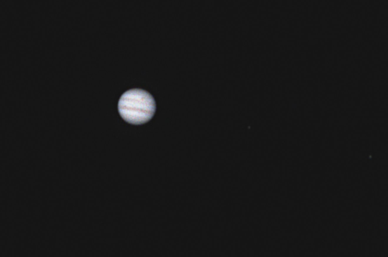 A blurry, low-resolution image shows the planet Jupiter as a pale, striped sphere against a black background, with two faint…