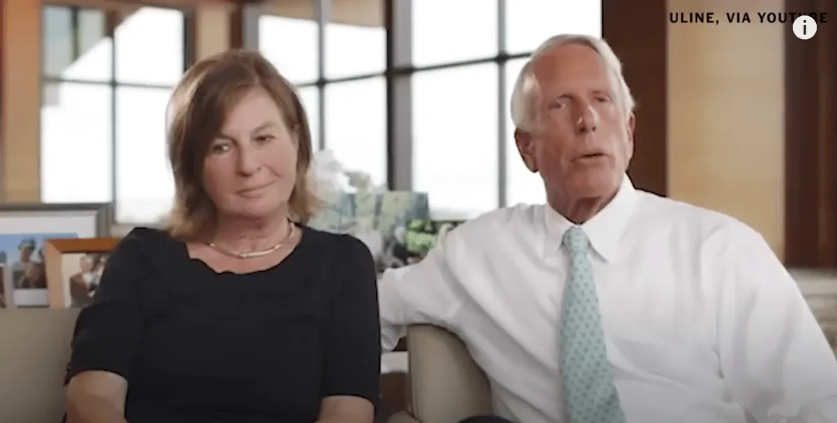 An older man in a white shirt and light blue tie sits beside a woman in a black top, both looking forward in what appears to…