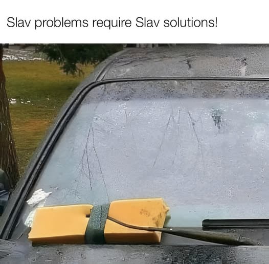 A close-up of a car's windshield covered in frost or ice, with a yellow sponge attached to the wiper arm by a green strap…