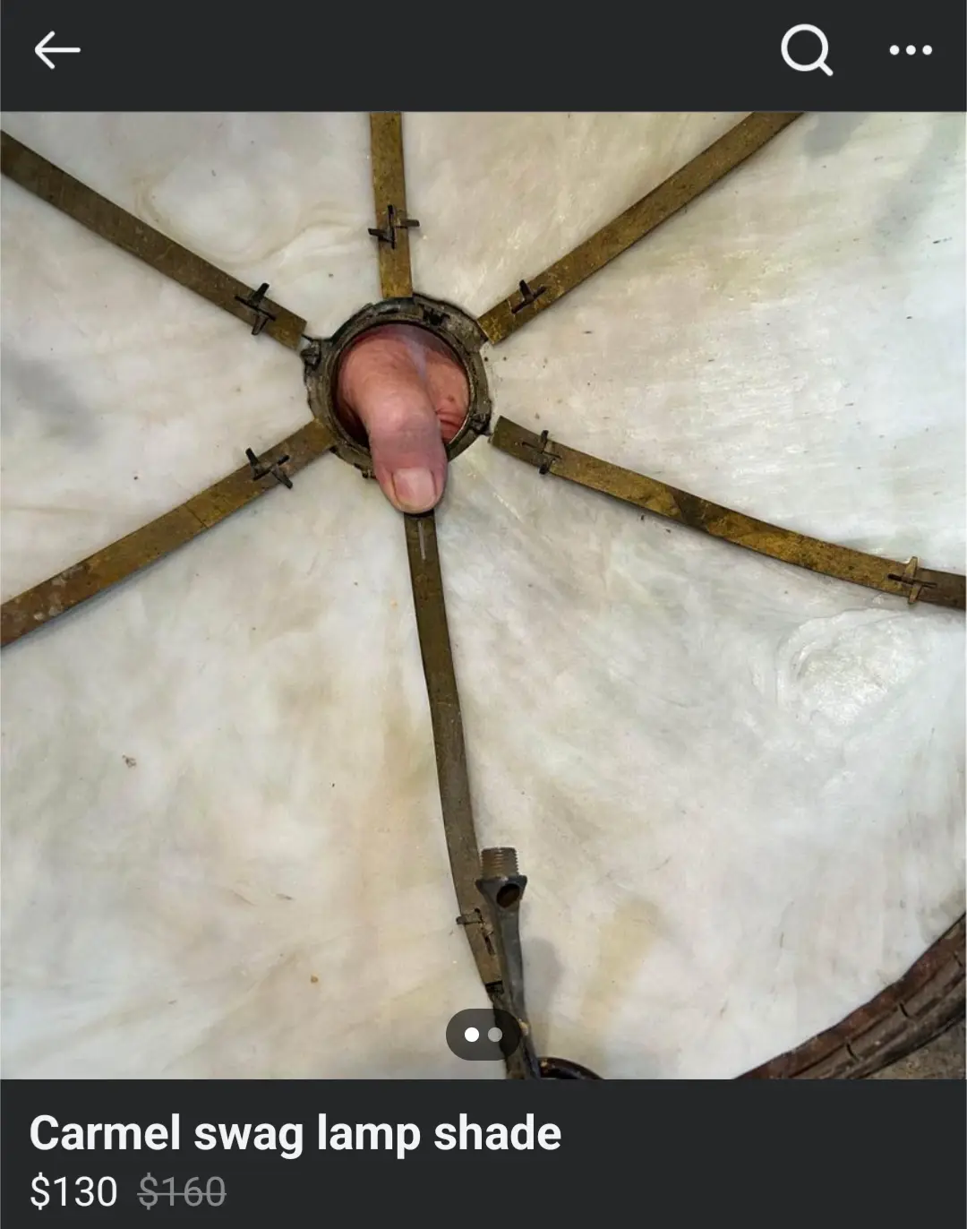A close-up image shows a person's thumb protruding from the center of a vintage-style lampshade, which has a white fabric…