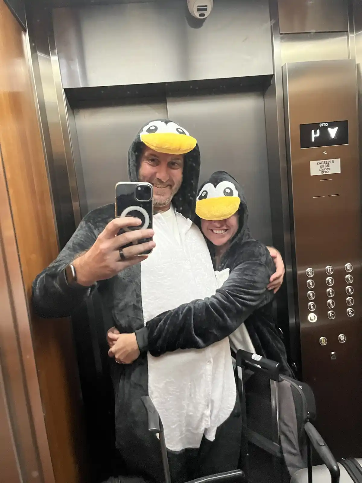 Two people in matching penguin hoodies take a selfie in an elevator, with the man holding the phone and the woman hugging…