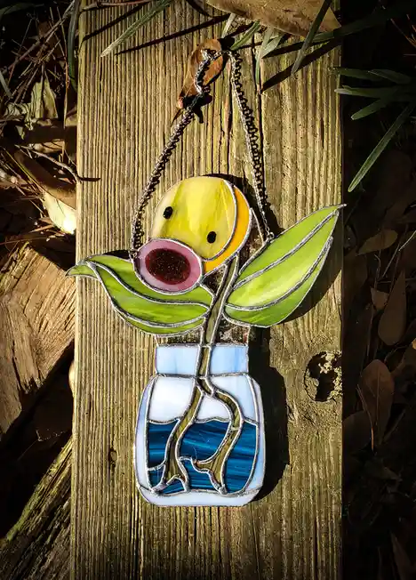 A stained glass pendant depicting a yellow bird with a large pink beak and green leaves, hanging from a chain on a wooden…