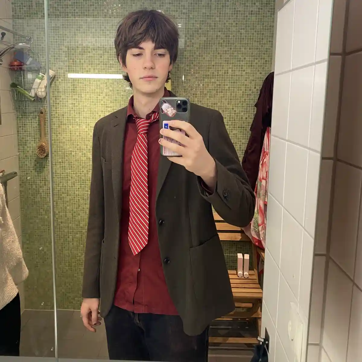 A young man with brown hair takes a mirror selfie in a bathroom wearing a brown blazer, red shirt, and a red-and-white…