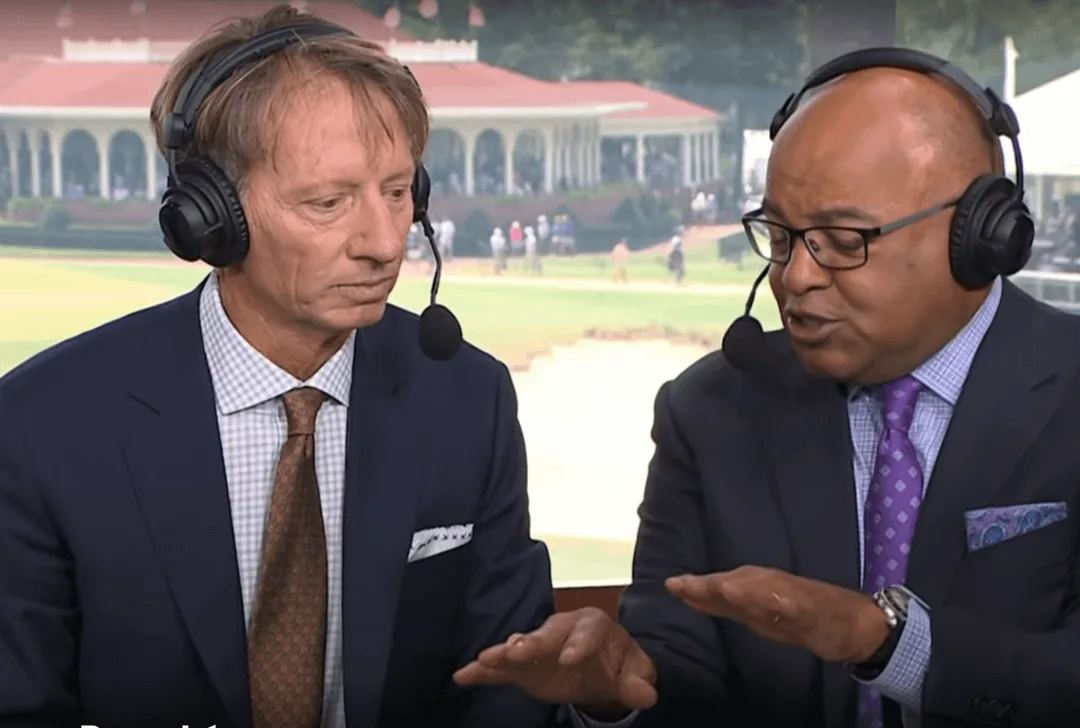 Two male sports commentators in suits and headsets sit in a broadcast booth with a golf course visible behind them. The man…