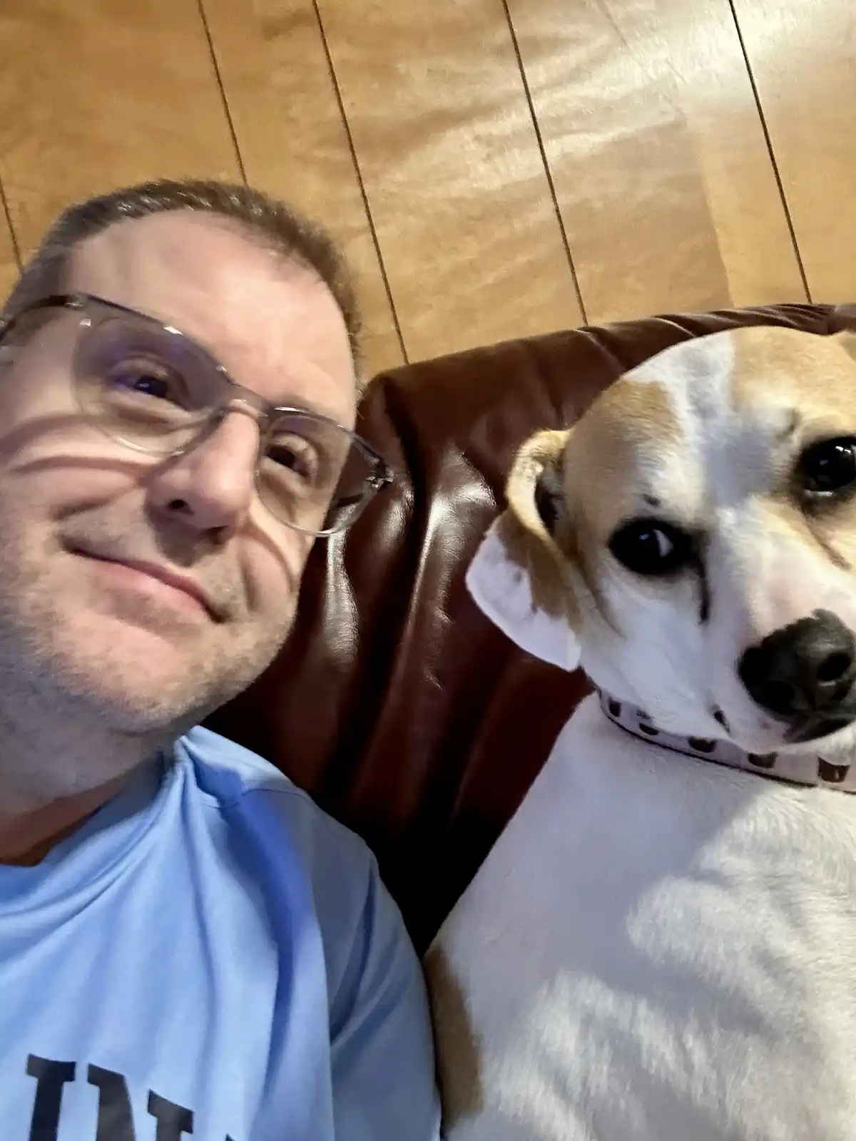 A man wearing glasses and a light blue t-shirt sits on a brown leather couch next to a white and tan dog with dark eyes and…