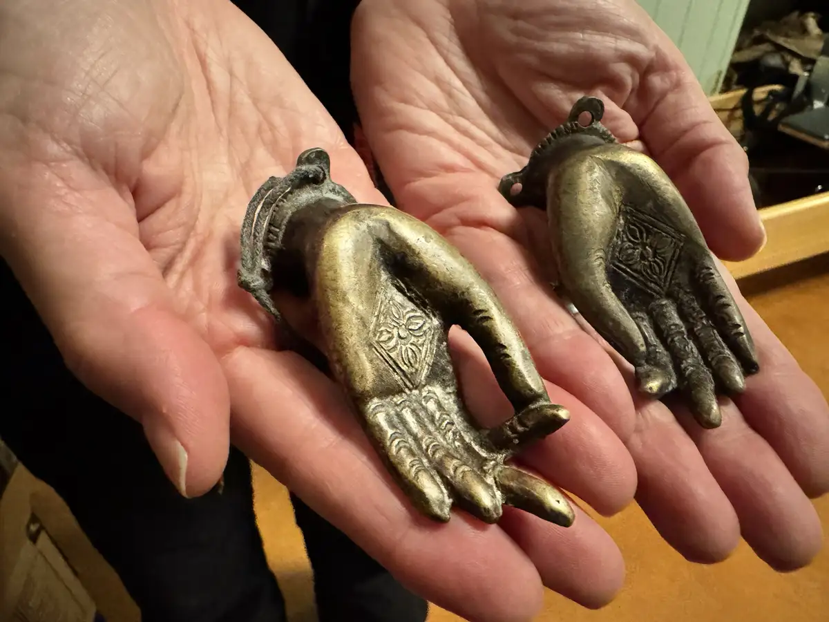 Two small, ornate bronze Buddha hand charms are held in a person's open palms. Each hand has intricate embossed patterns on…