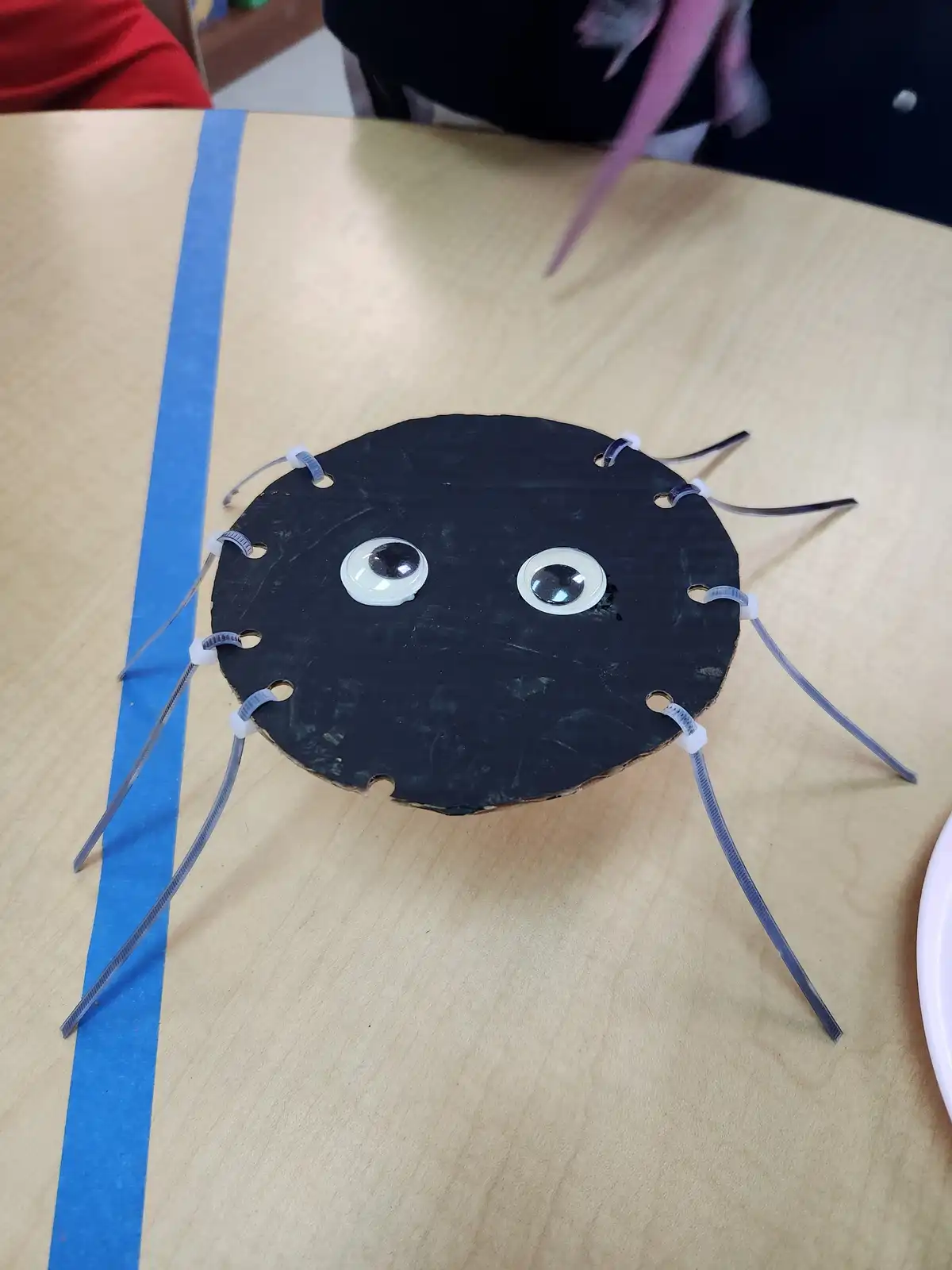 A handmade spider craft sits on a wooden table, constructed from a black circular disc with googly eyes and six legs made…